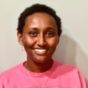 Sara Sirag smiles at the camera in front of a while wall. She's wearing a pink long sleeve shirt.